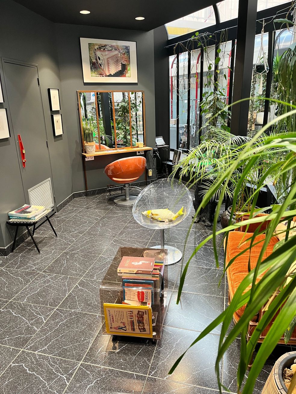 Un salon moderne avec un plafond sombre, un sol en terrazzo, un fauteuil de coiffure rouge et de luxuriantes plantes d'intérieur près de grandes fenêtres.