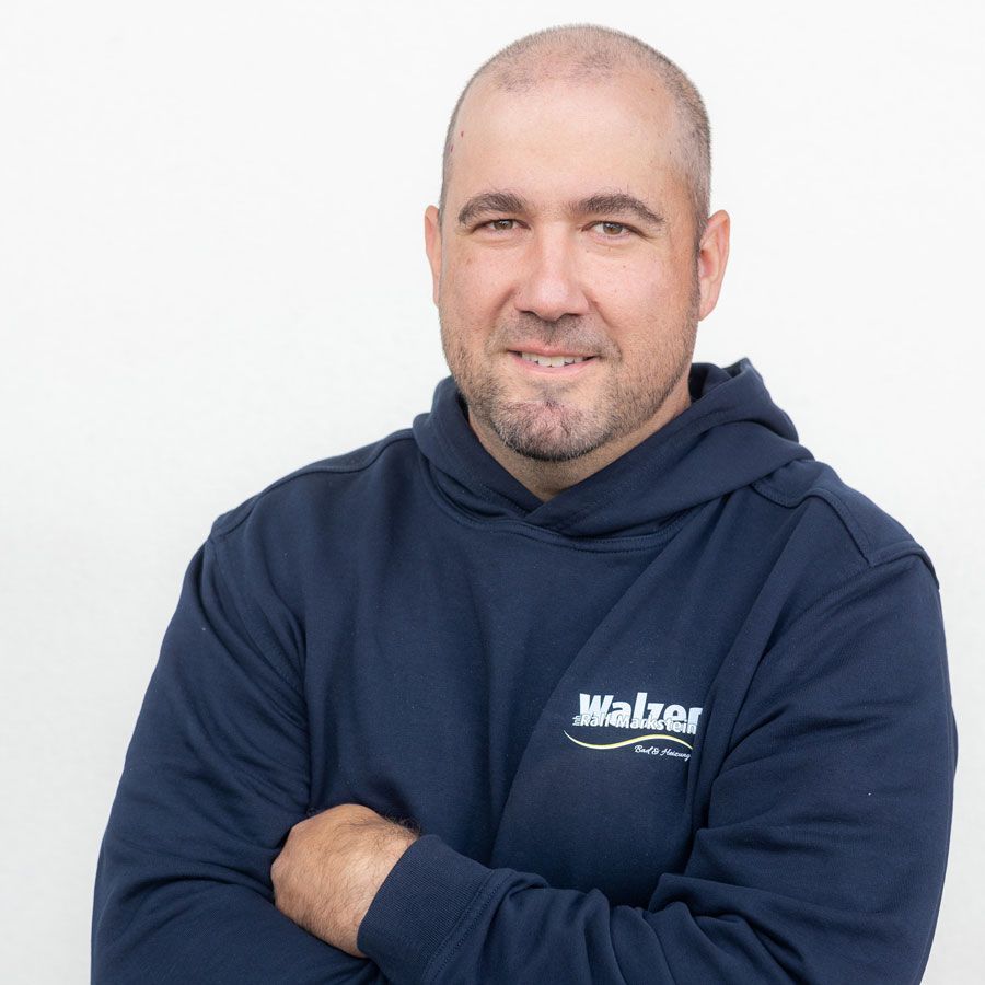 Ein Mann in einem gelben Poloshirt steht im Freien, lächelt und hat die Arme verschränkt.