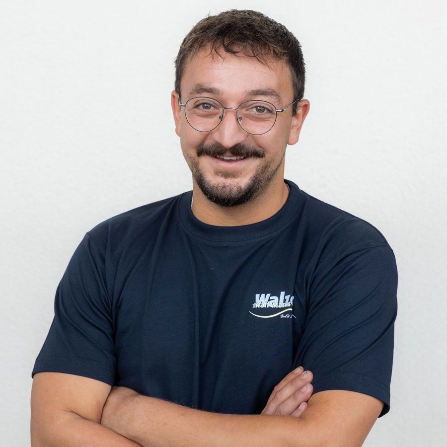 Lächelnde Person in gelbem Poloshirt und Brille steht mit verschränkten Armen in einem Innenraum.
