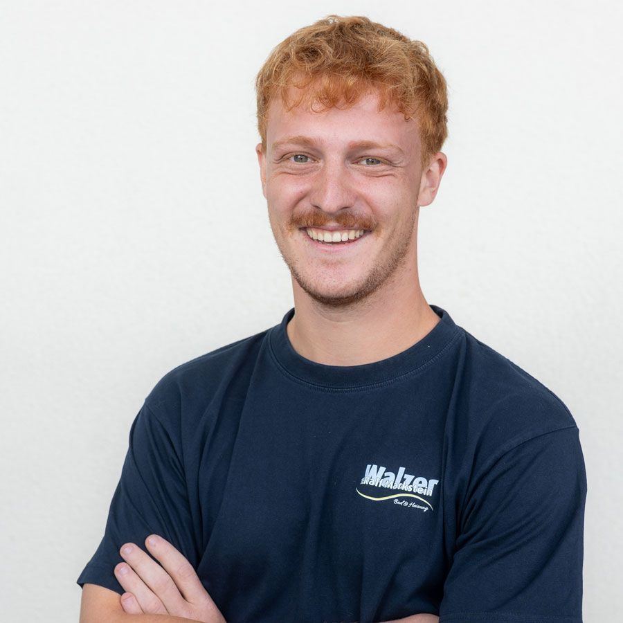 Eine lächelnde Person in einem leuchtend gelben Poloshirt steht drinnen in der Nähe eines Fensters.