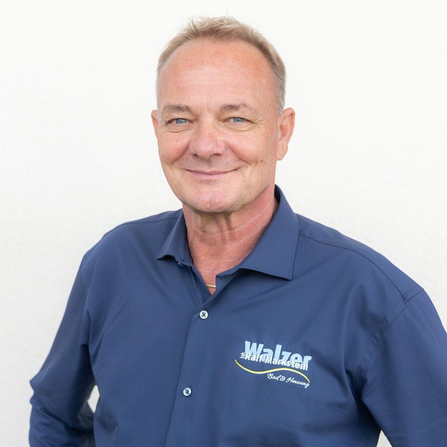 Ein lächelnder Mann in einem gelben Poloshirt steht mit verschränkten Armen an einer beigen Wand.
