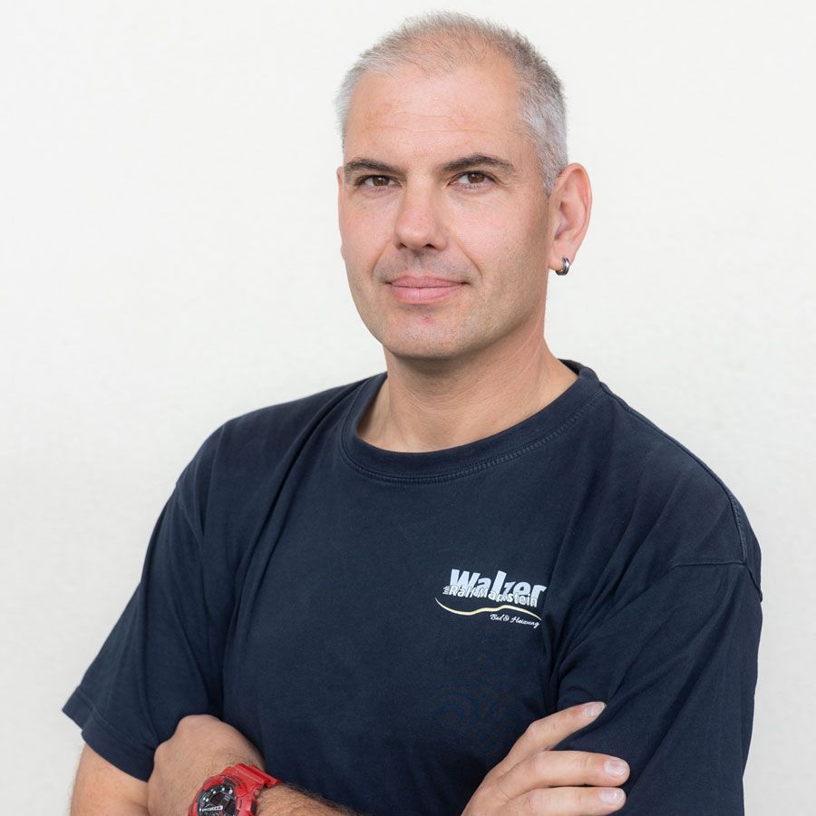 Mann in gelbem Poloshirt, der mit verschränkten Armen an einer beigen Wand steht.