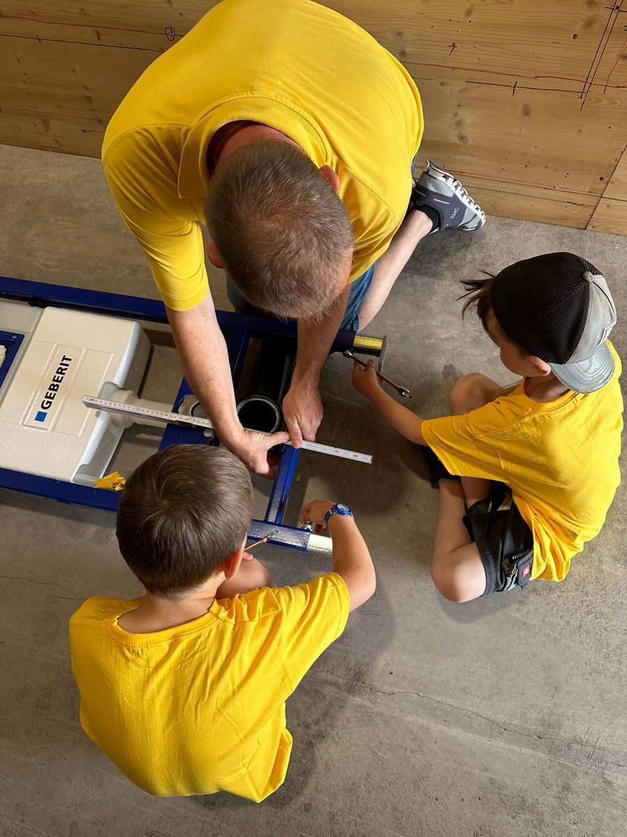 Drei Personen in gelben Hemden untersuchen ein Tablet, das neben Kisten und Werkzeugen auf dem Boden liegt.