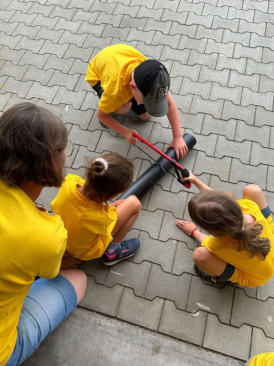 Drei Kinder und ein Erwachsener spielen auf einem Backsteinweg Tauziehen mit einem schwarzen Gartenschlauch.