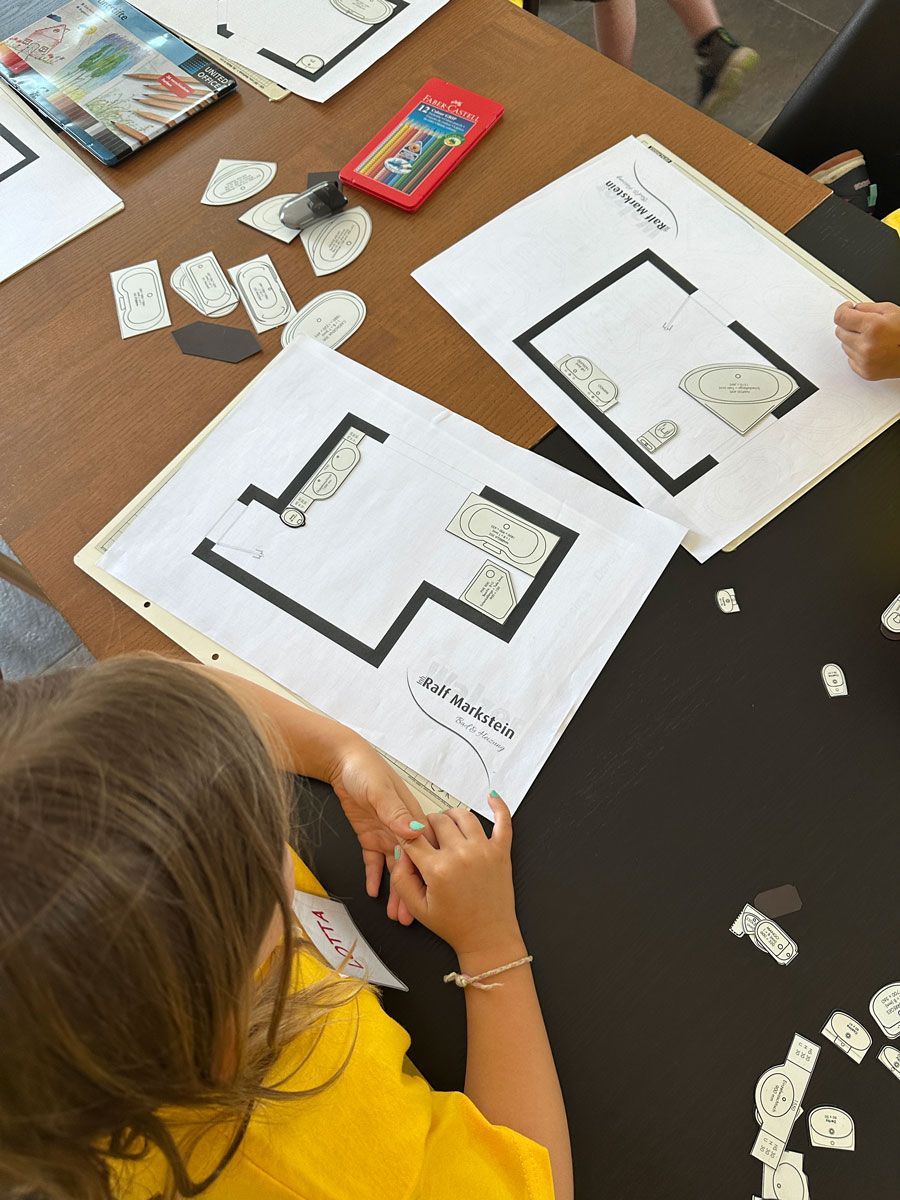 Ein Kind bearbeitet an einem Tisch mit Stiften und kleinen Papierstücken Arbeitsblätter mit Grundrissen.
