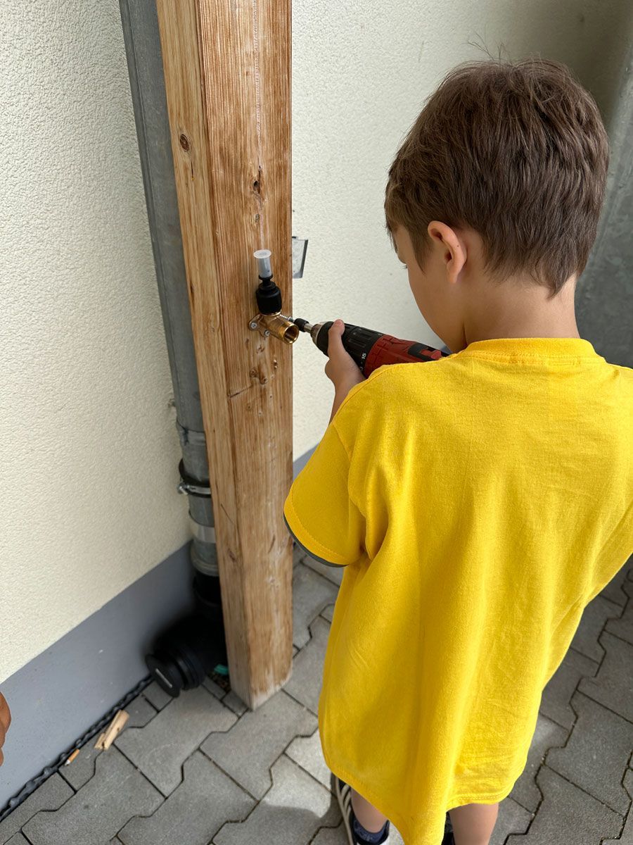 Kind in gelbem Hemd benutzt ein Werkzeug, um an einem Holzpfosten an einer Mauer zu arbeiten.