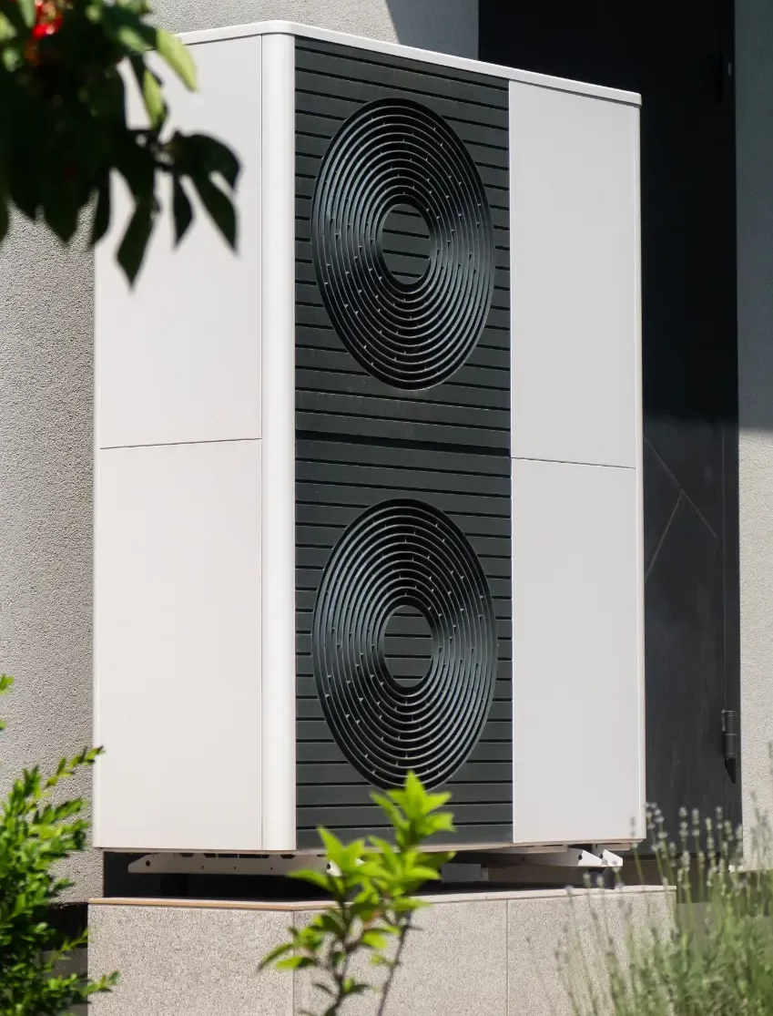 Außenliegende Wärmepumpeneinheit mit zwei runden Ventilatoren, weiß und schwarz, neben einem Gebäude.