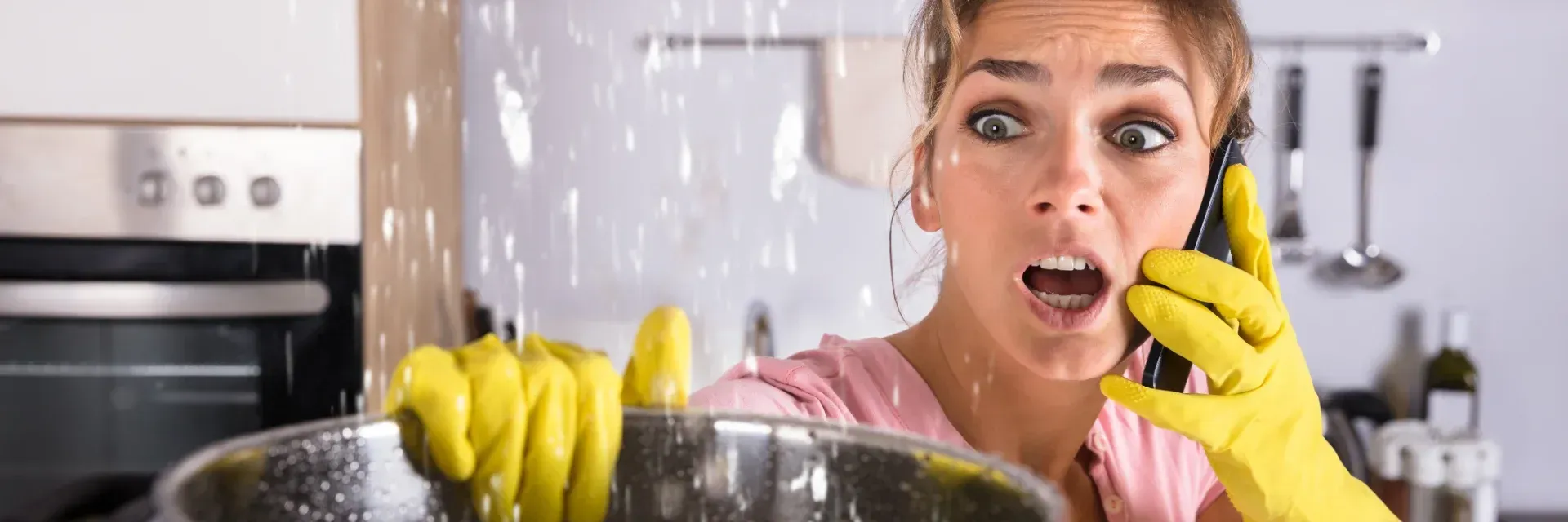 Eine Frau in der Küche, die mit behandschuhten Händen telefoniert, ist schockiert, als Wasser über ein Spülbecken voller gelber Putzhandschuhe schwappt.