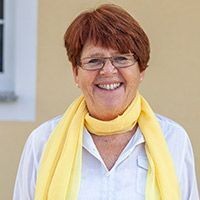 Eine lächelnde Person mit Brille, gelbem Schal und weißem Hemd steht im Freien vor einer beigen Wand.