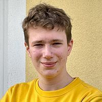 Person in gelbem Hemd lächelt im Freien vor einer strukturierten Wand.