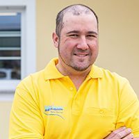 Ein Mann in einem gelben Poloshirt steht im Freien, lächelt und hat die Arme verschränkt.