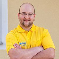Mann in einem gelben Poloshirt mit verschränkten Armen, der vor einer beigen Wand steht.
