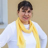 Person lächelt in weißem Oberteil und gelbem Schal vor beiger Wand