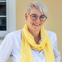 Lächelnde Person mit kurzen grauen Haaren, runder Brille, weißem Hemd und gelbem Schal vor einer beigen Wand