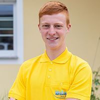 Eine lächelnde Person in einem leuchtend gelben Poloshirt steht drinnen in der Nähe eines Fensters.