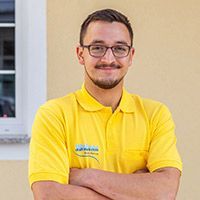 Lächelnde Person in gelbem Poloshirt und Brille steht mit verschränkten Armen in einem Innenraum.