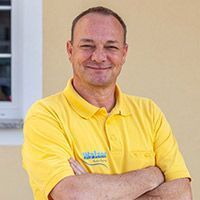 Ein lächelnder Mann in einem gelben Poloshirt steht mit verschränkten Armen an einer beigen Wand.