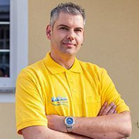 Mann in gelbem Poloshirt, der mit verschränkten Armen an einer beigen Wand steht.