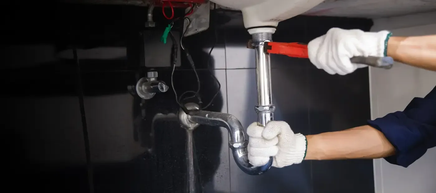 Mit behandschuhten Händen wird mithilfe eines Schraubenschlüssels die Rohrleitung unter einem Waschbecken festgezogen, wobei rote und schwarze Rohre sichtbar sind.