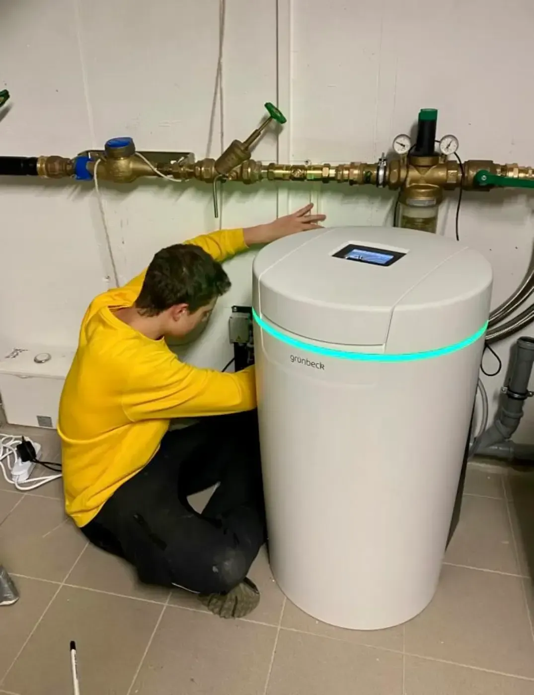 Eine Person kniet neben einem weißen Warmwasserbereiter in einem Hauswirtschaftsraum mit freiliegenden Rohren.