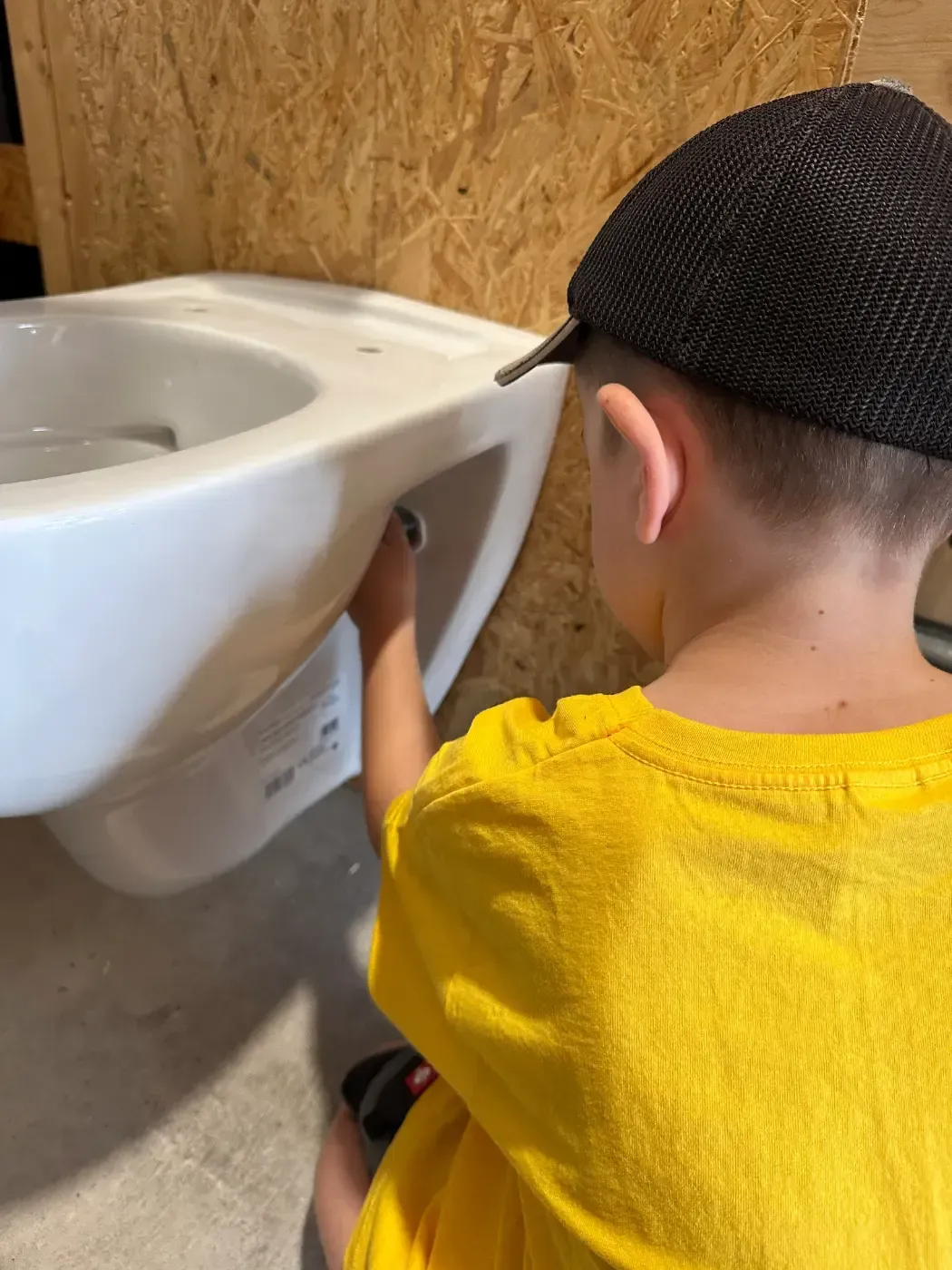 Kind in gelbem Hemd und schwarzer Mütze reinigt eine weiße Toilette in einer Toilettenkabine.