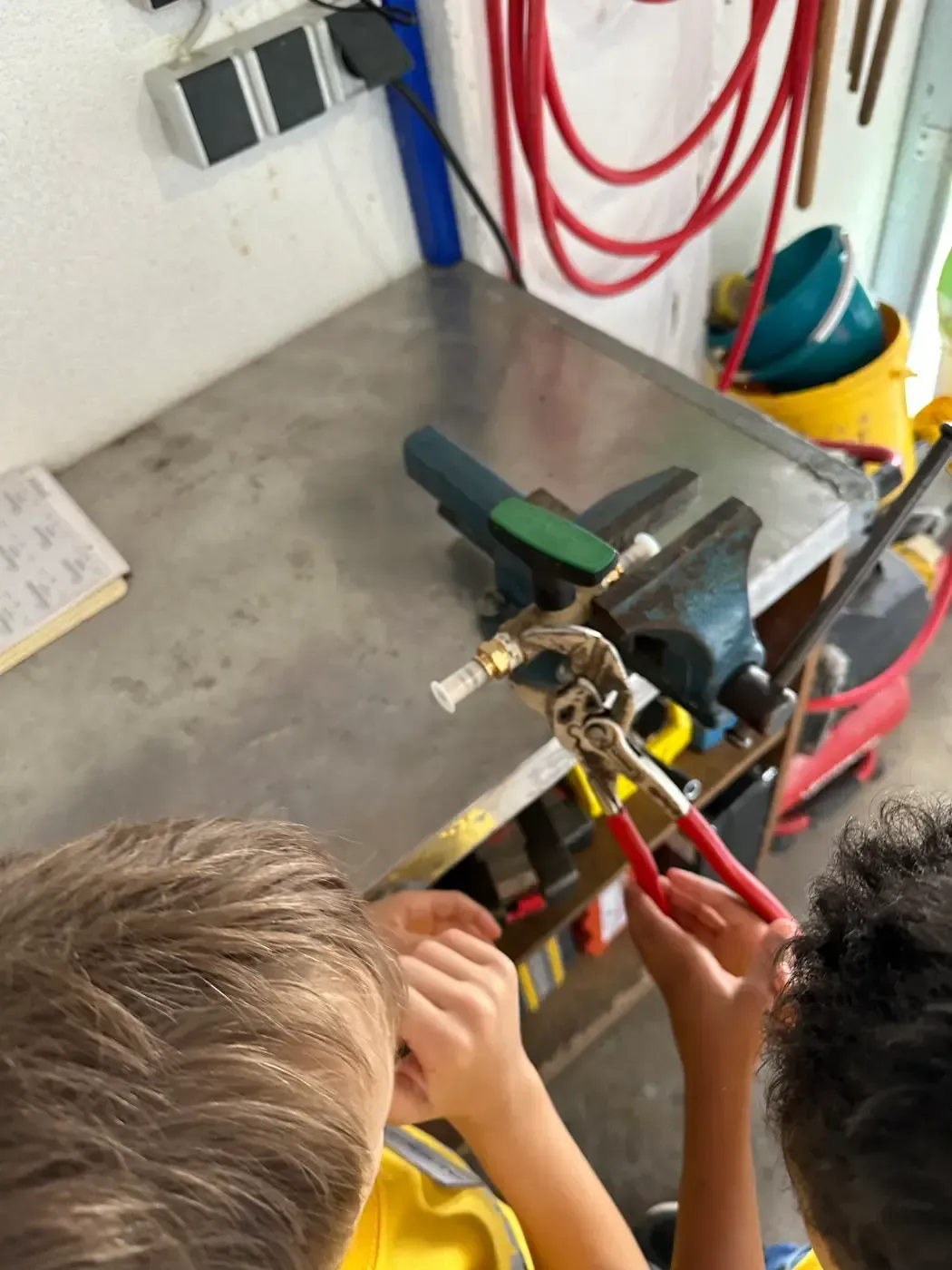 Zwei Kinder benutzen in einer Werkstatt an einer Metallwerkbank eine Zange, um an einem kleinen Gerät zu arbeiten.