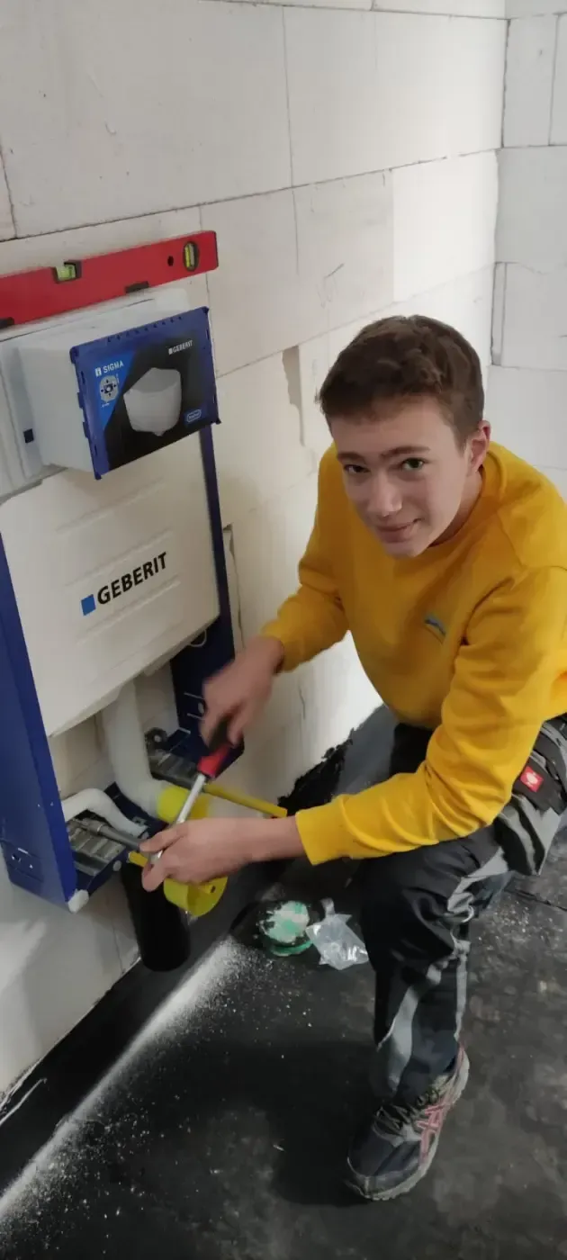 Eine Person in einem gelben Sweatshirt hockt neben einer an der Wand montierten Maschine und hantiert in einem gefliesten Raum mit bunten Kabeln.