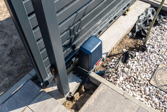 Moteur de portail à côté d'un portail coulissant en métal sur une allée en gravier et en béton.