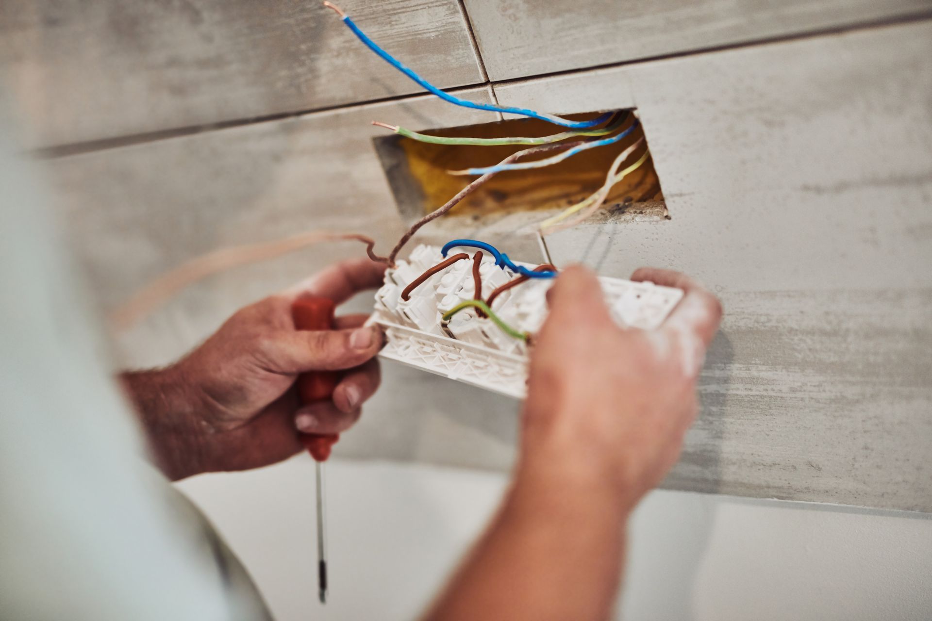 Des mains câblent une prise électrique dans une boîte murale, les câbles étant apparents.