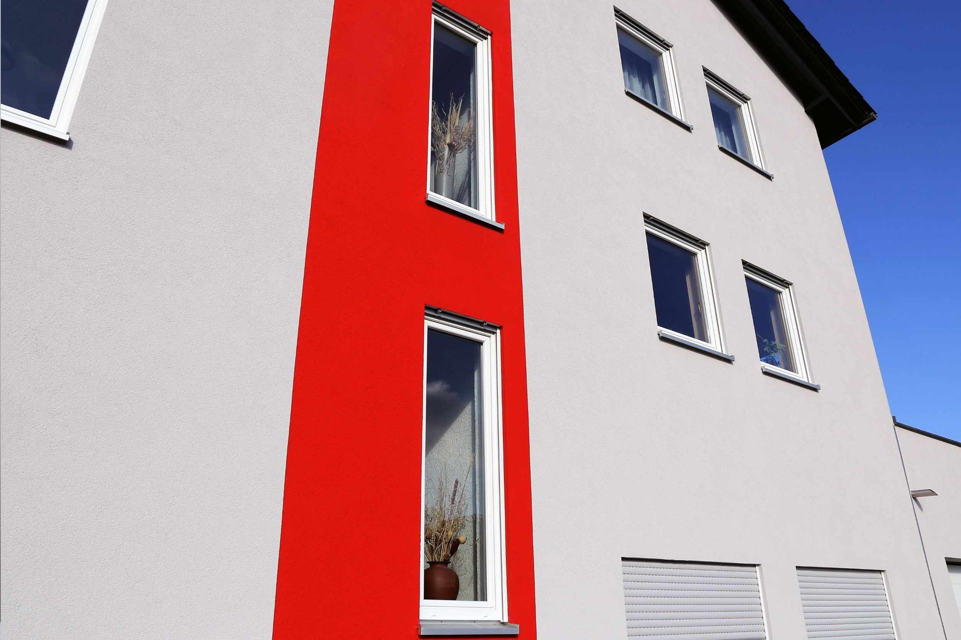 Exterior de un edificio moderno con fachada gris y franja roja. Ventanas enmarcadas en blanco bajo un cielo azul claro.