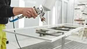 Persona pintando con aerosol un marco de puerta blanco con una pistola en un taller muy iluminado.