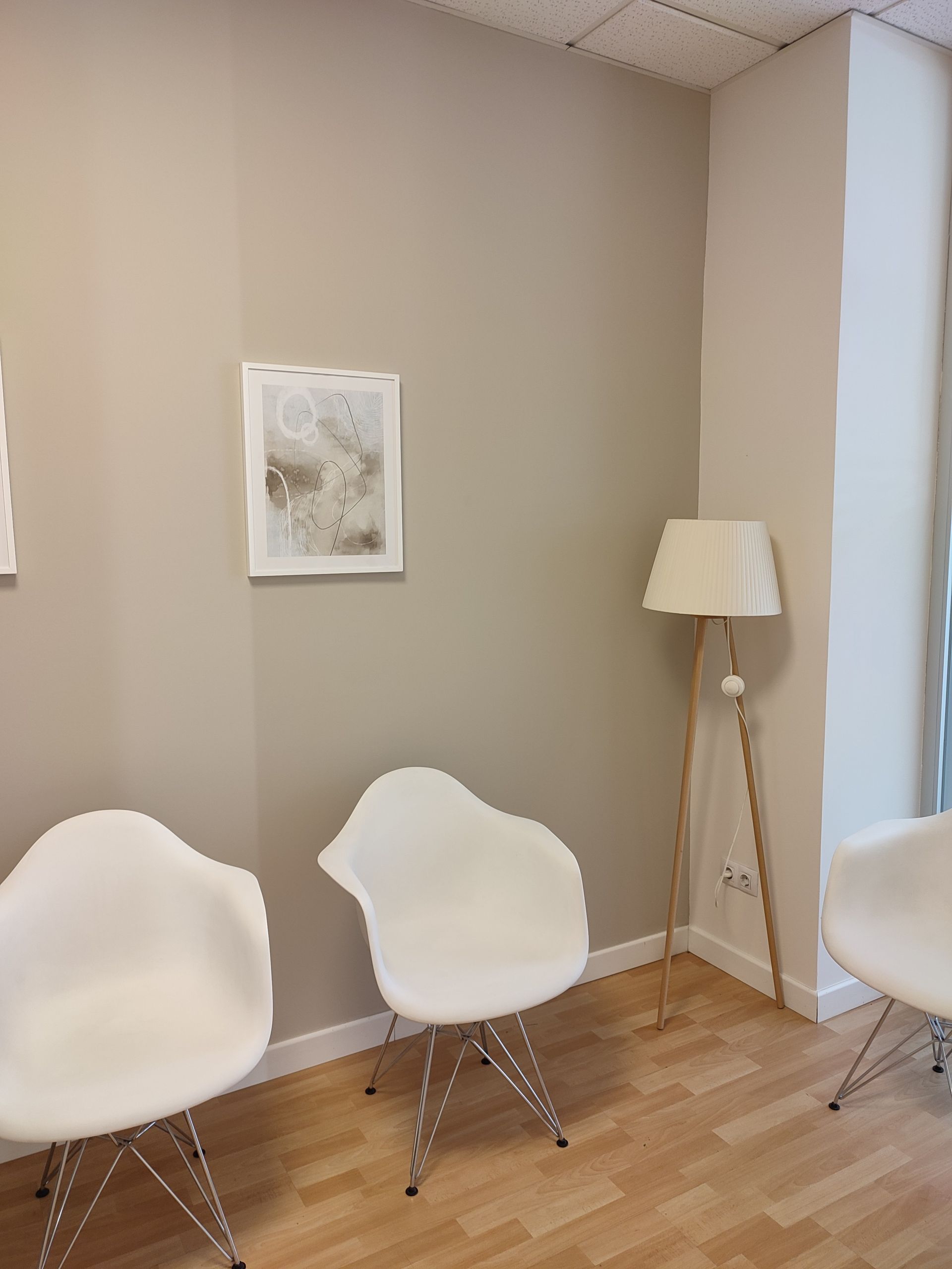 Una sala de espera con sillas blancas, una lámpara de pie y obras de arte en una pared beige.
