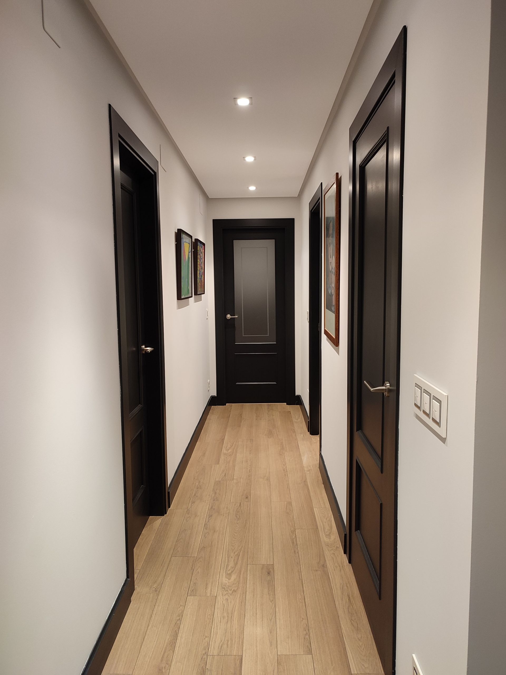 Un largo pasillo con suelos de madera clara, paredes blancas y puertas marrón oscuro. Iluminación empotrada ilumina el espacio.