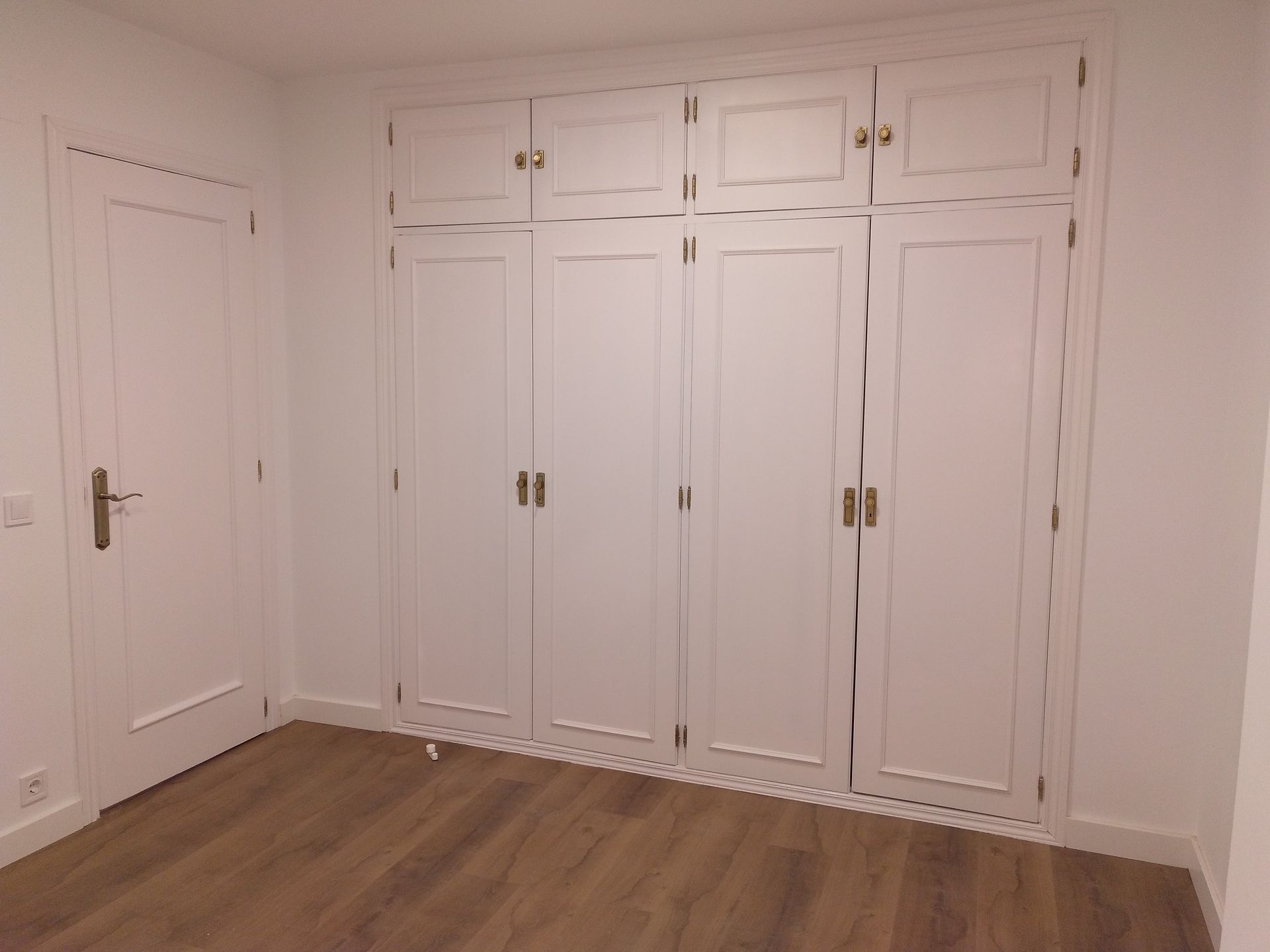 Armario empotrado blanco de seis puertas y tiradores dorados en un dormitorio con suelo de madera y puerta blanca.