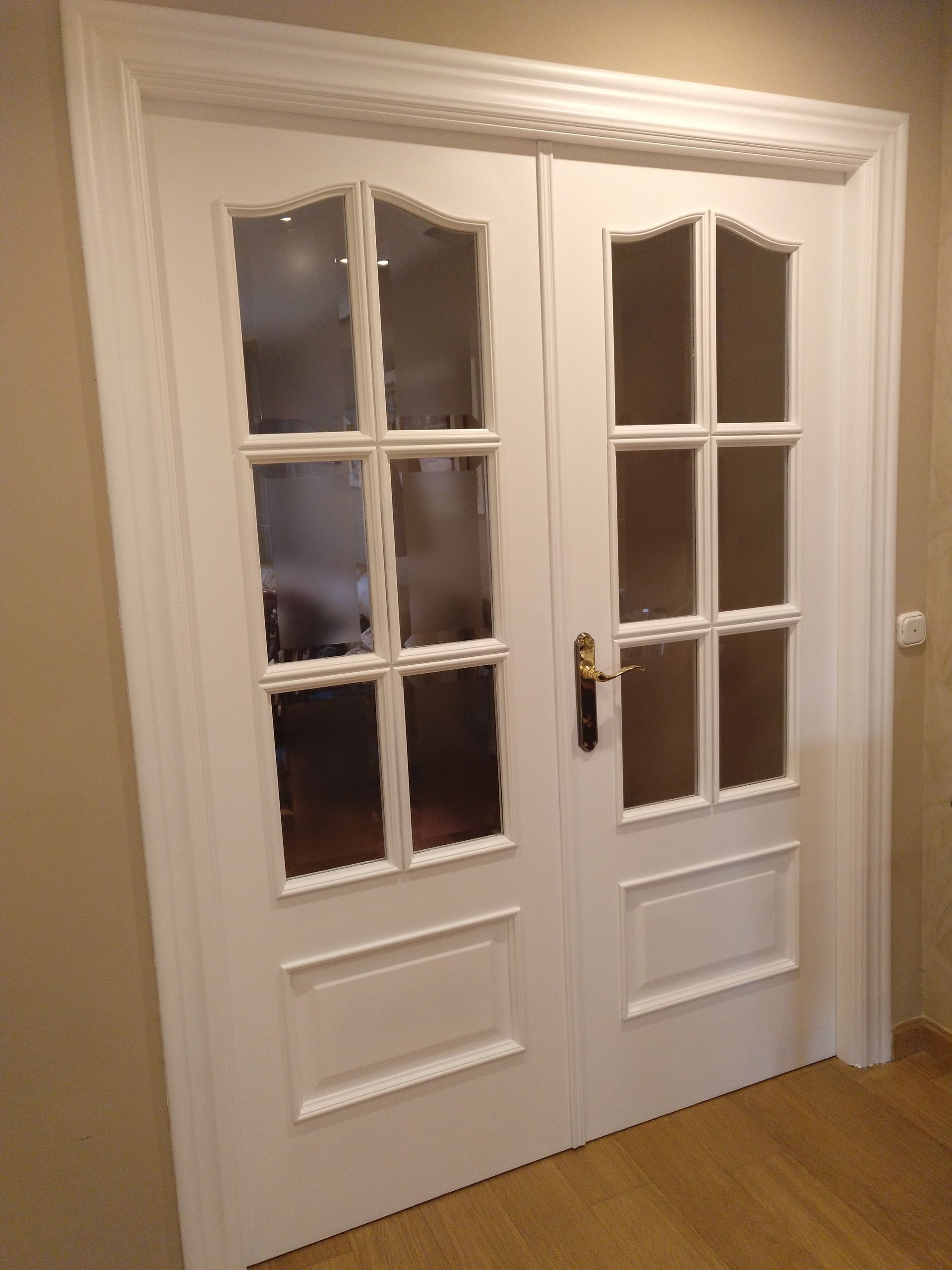 Puertas dobles blancas con paneles de vidrio, con moldura decorativa y tirador dorado, en un pasillo de color claro.