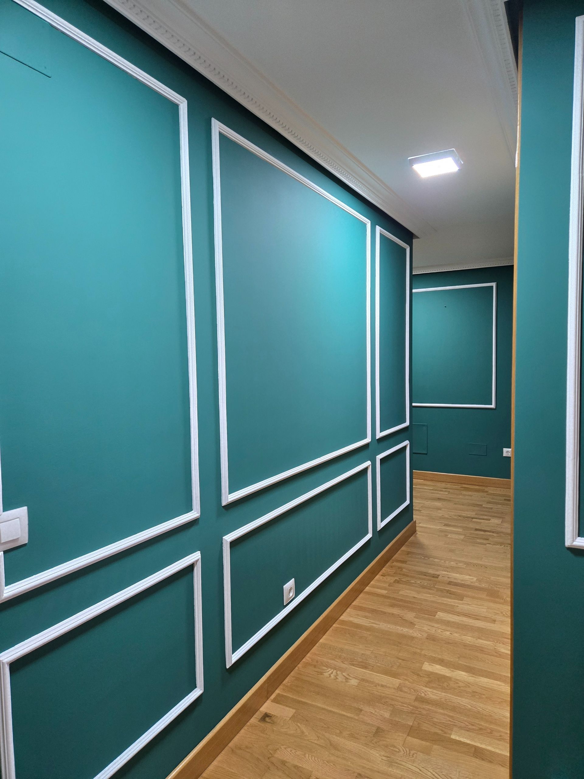 Pasillo verde azulado con paneles de pared con marco blanco y suelo de madera. Al fondo se ve una habitación rectangular.