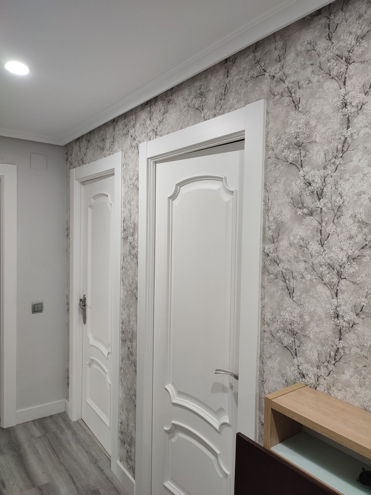 Pasillo con puertas y molduras blancas, papel tapiz con estampado gris y una mesa auxiliar de madera.