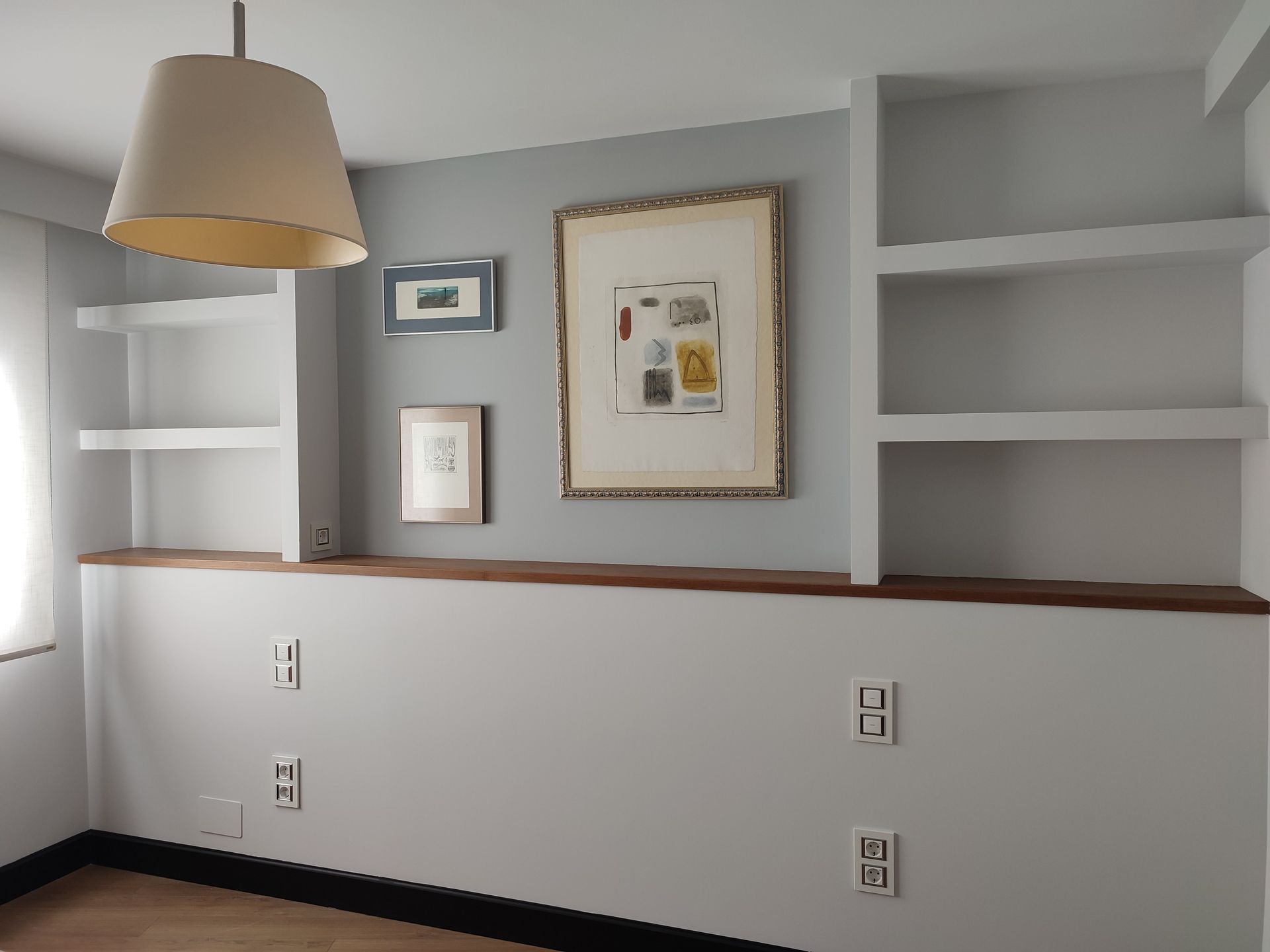 Estantes empotrados y obras de arte sobre una repisa de madera en una pared gris. Una lámpara cuelga del techo.