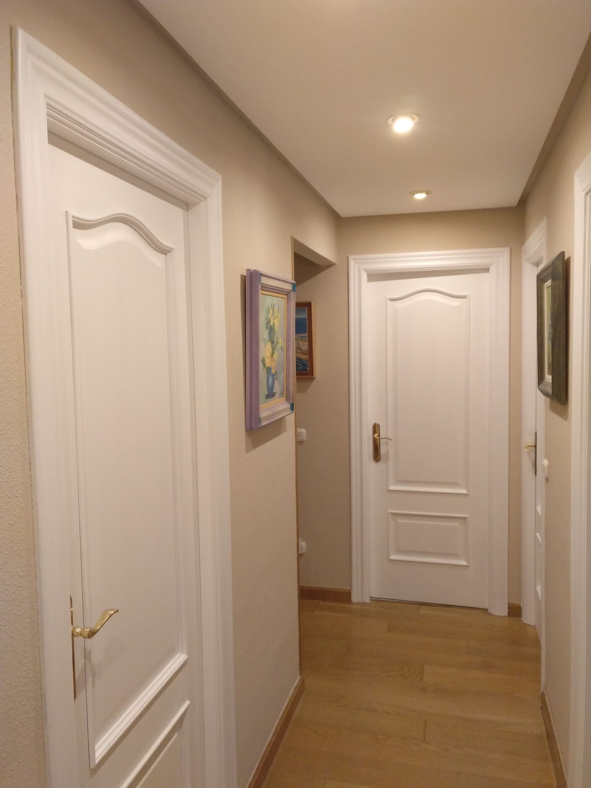 Pasillo con paredes de color beige claro, puertas blancas y suelo de madera; dos cuadros cuelgan en la pared.