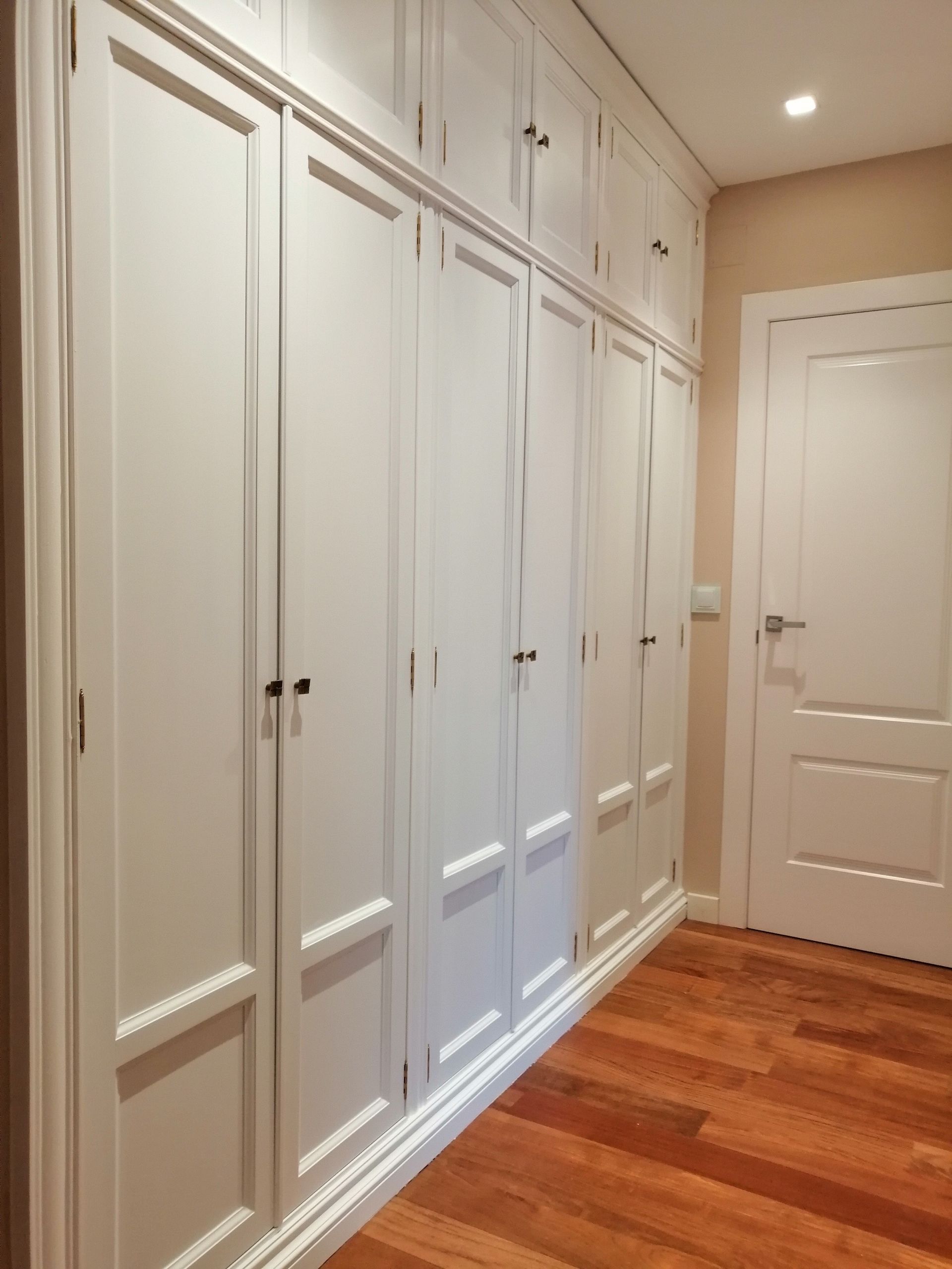 Un armario empotrado largo, de color blanco, con múltiples puertas, que contrasta con un suelo de madera y una puerta en el lado derecho.