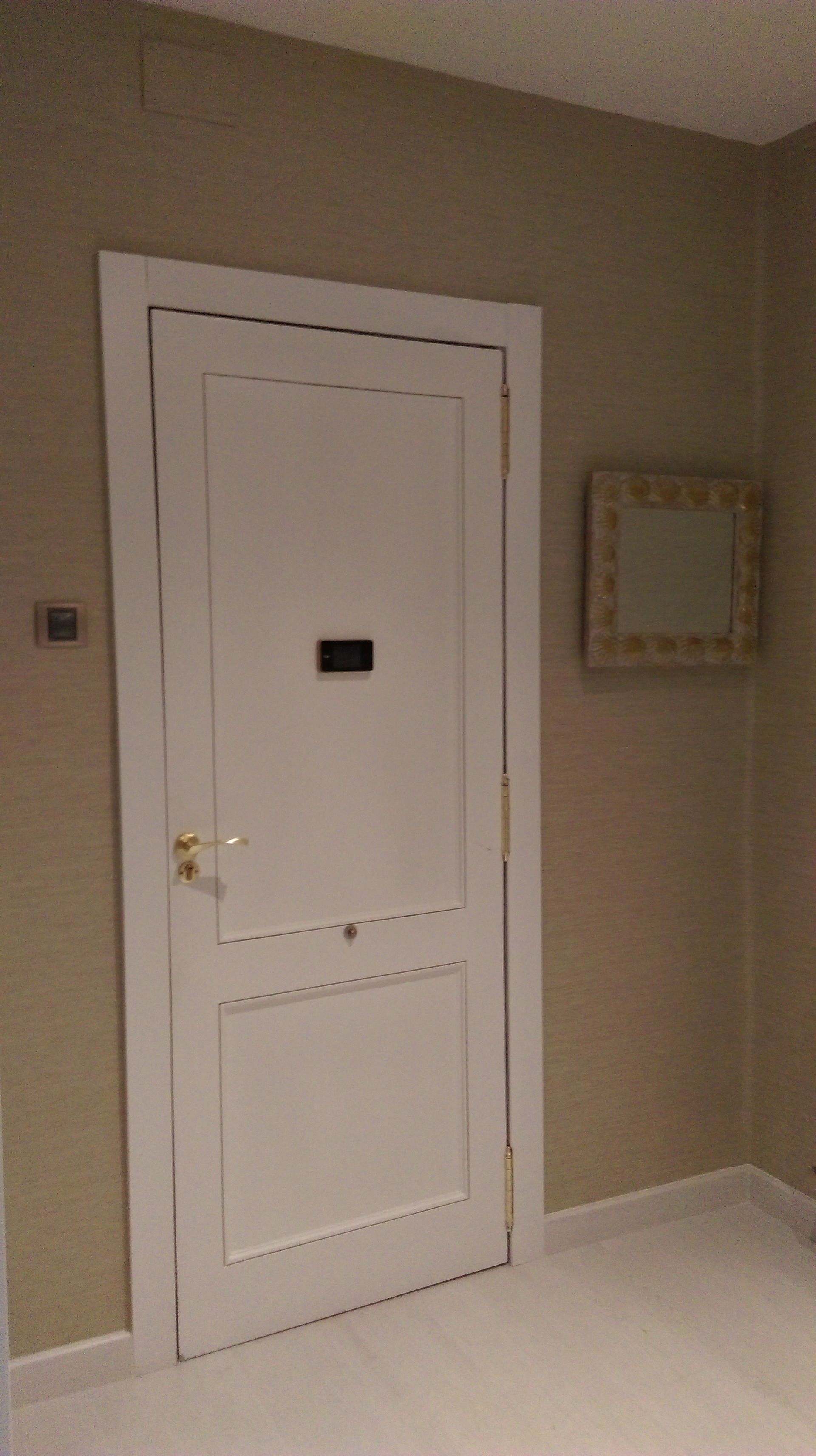 Puerta blanca con un pequeño letrero negro, tirador dorado y un marco decorativo en un pasillo beige. Un pequeño espejo cuelga a la derecha.