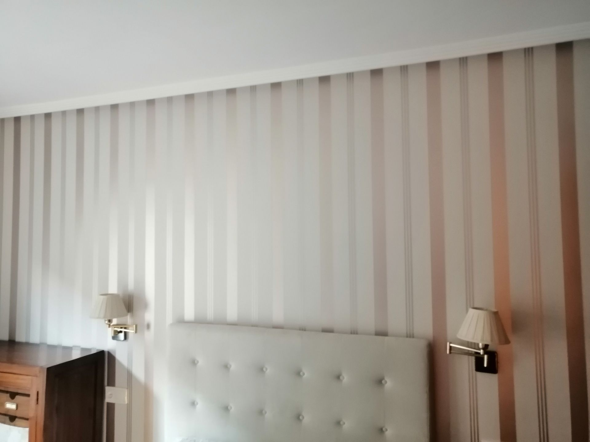 Pared del dormitorio con papel pintado de rayas verticales en tonos blancos y cobre, sobre un cabecero acolchado y una lámpara de aplique.