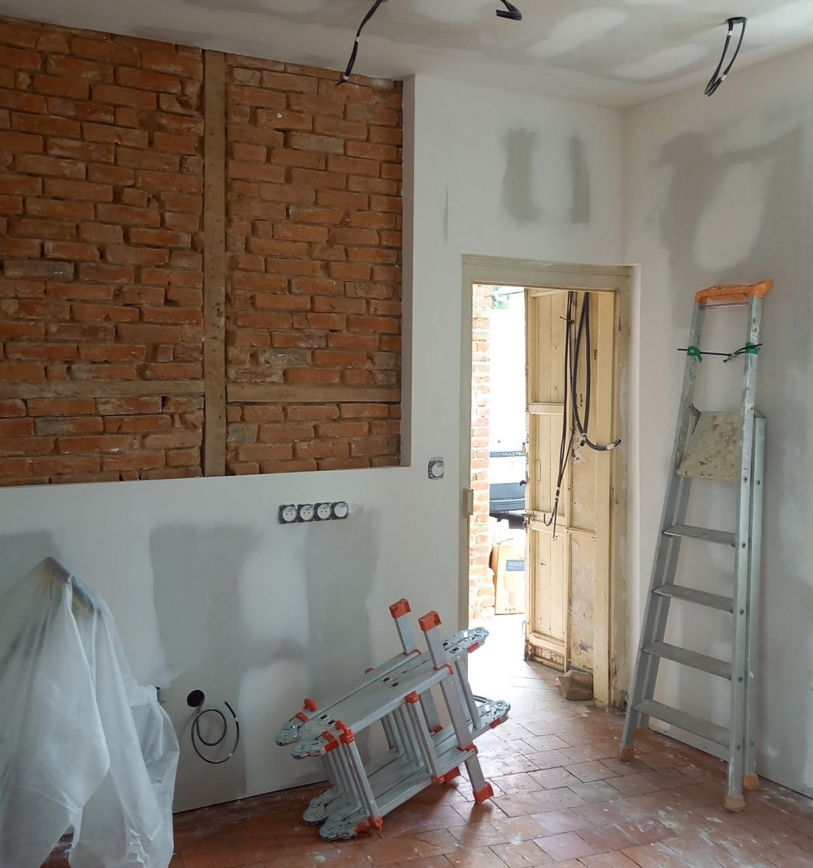 Rénovation de la pièce en cours ; mur de briques apparentes, cloisons sèches, échelles, porte ouverte.