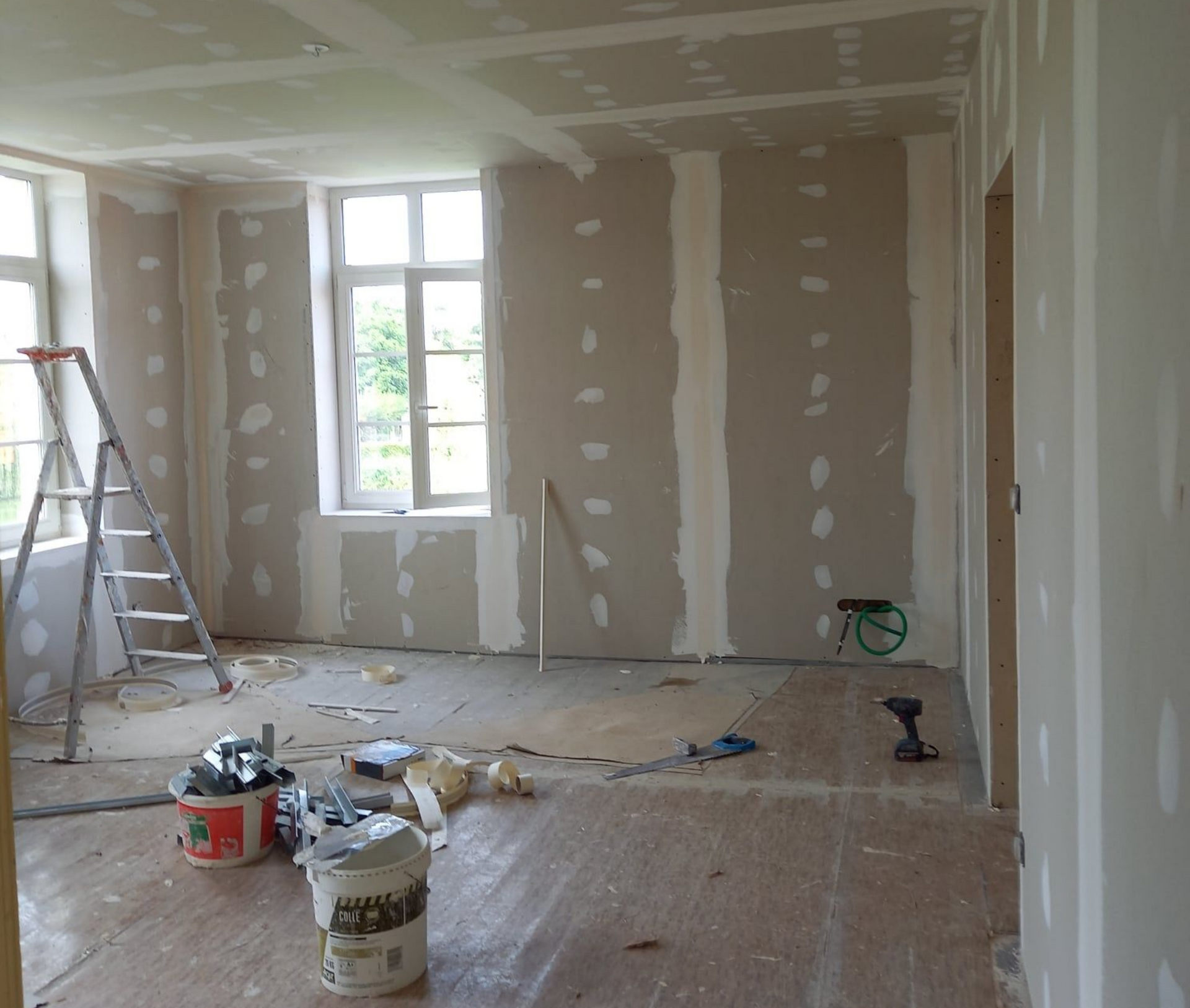 Intérieur d'une pièce en construction, avec des plaques de plâtre posées sur les murs. On aperçoit une échelle, des outils et des seaux.