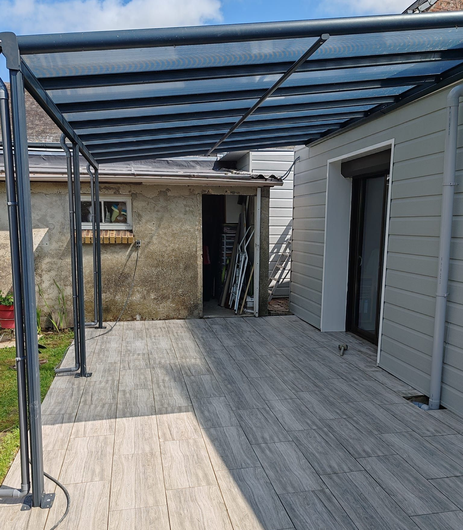 Une pergola grise recouvre une terrasse en bois jouxtant un bâtiment au bardage gris et à la structure en pierre patinée.