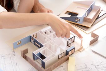 Une personne désigne du doigt une petite maquette architecturale d'une maison aux éléments bleu clair et en bois.