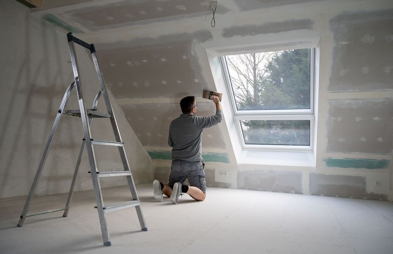 Une personne ponce des plaques de plâtre autour d'une fenêtre dans une pièce en construction, à l'aide d'une échelle.