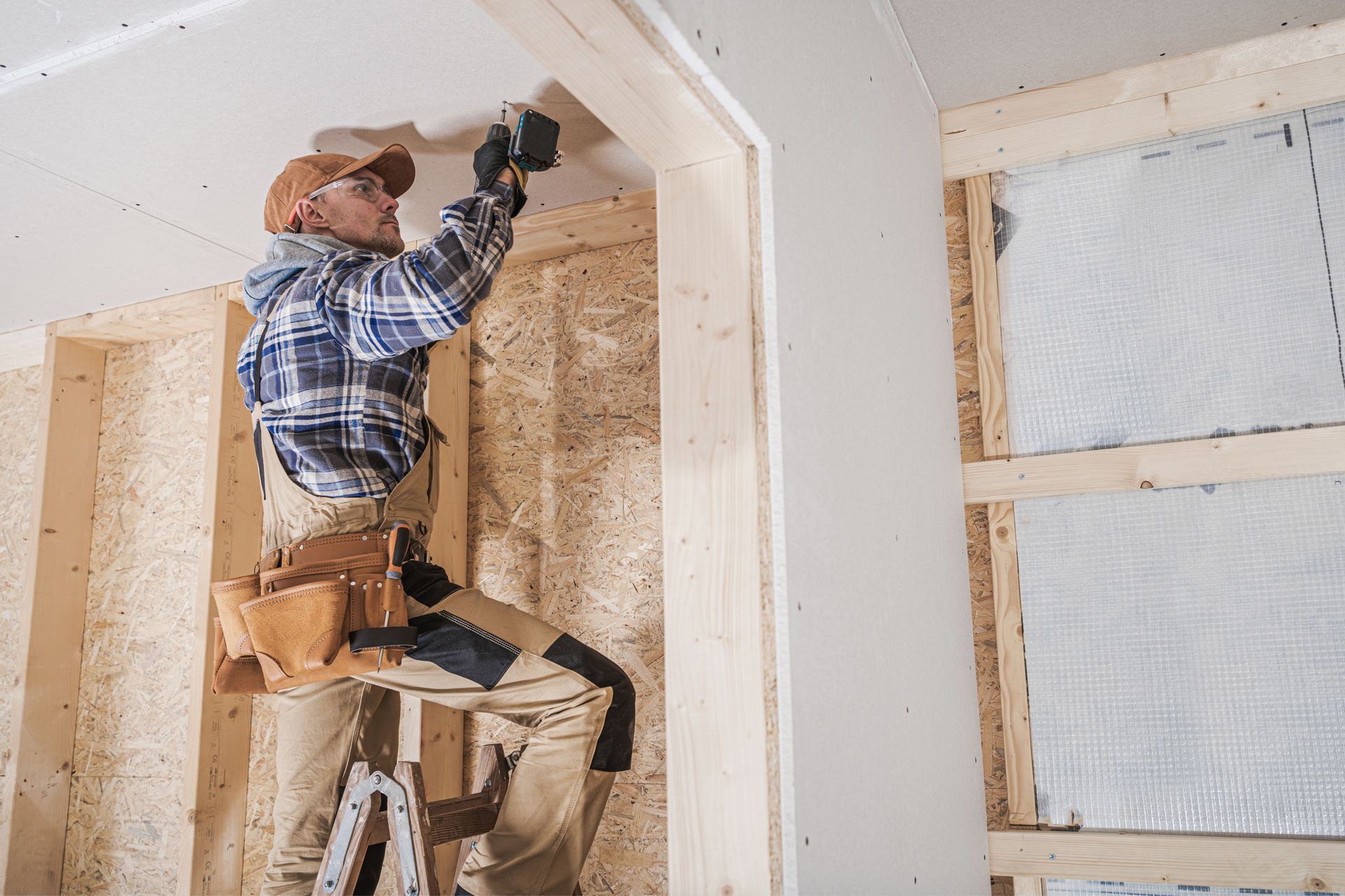 Un ouvrier du bâtiment, juché sur une échelle, installe des plaques de plâtre dans une pièce en construction ; la structure en bois est visible.