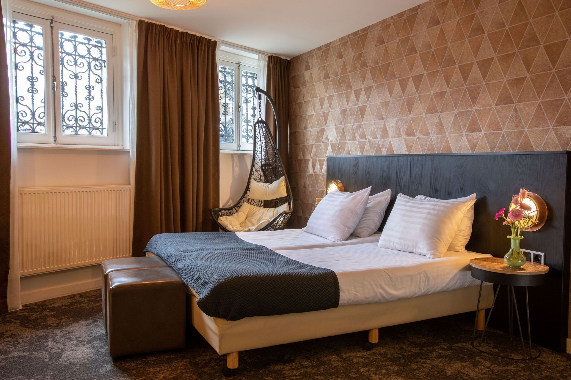 Slaapkamer met een tweepersoonsbed, hangstoel, bruine gordijnen en behang met patroon.