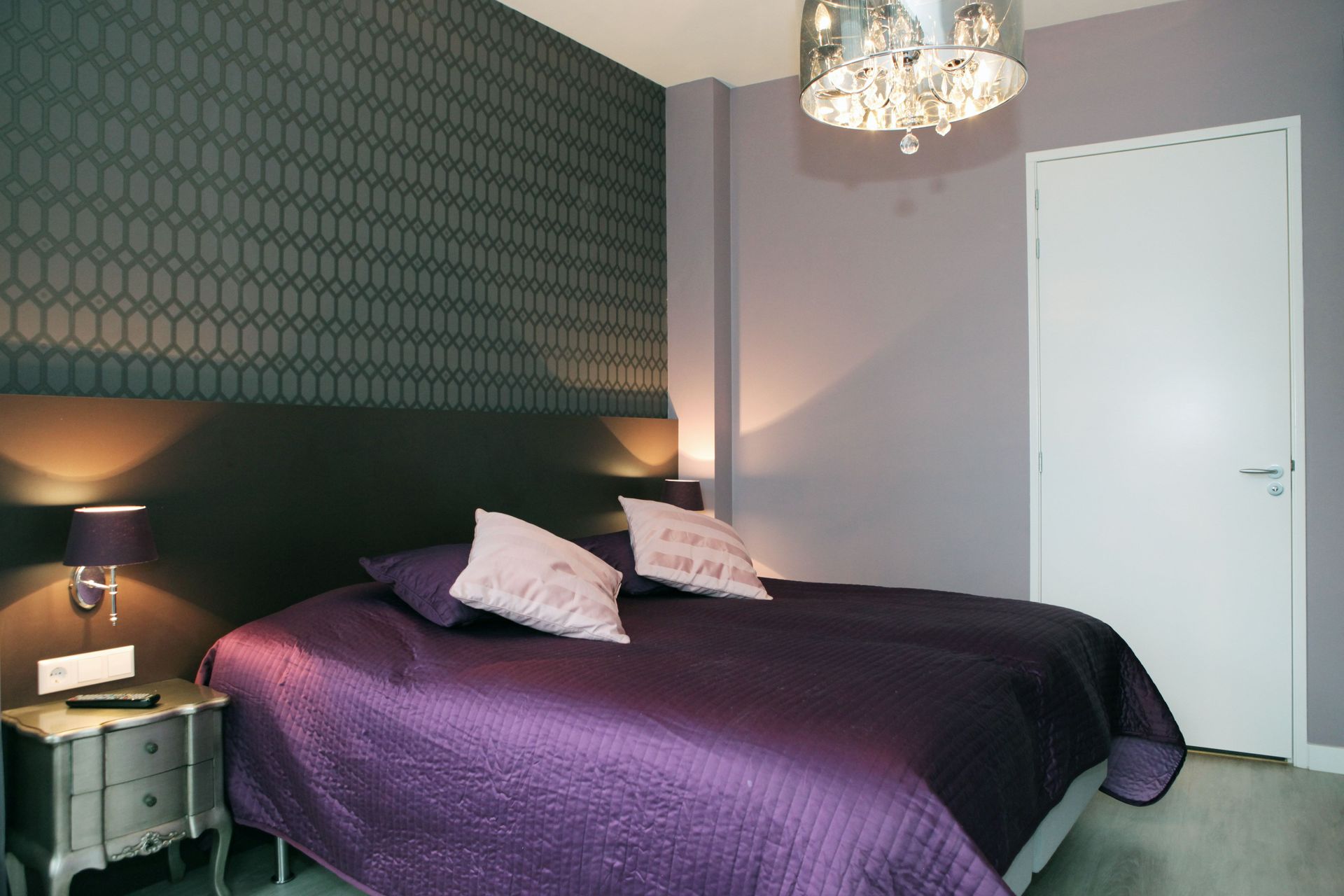 Slaapkamer met paars thema, behang met patroon, bed, nachtkastje, lampen en een decoratieve kroonluchter.